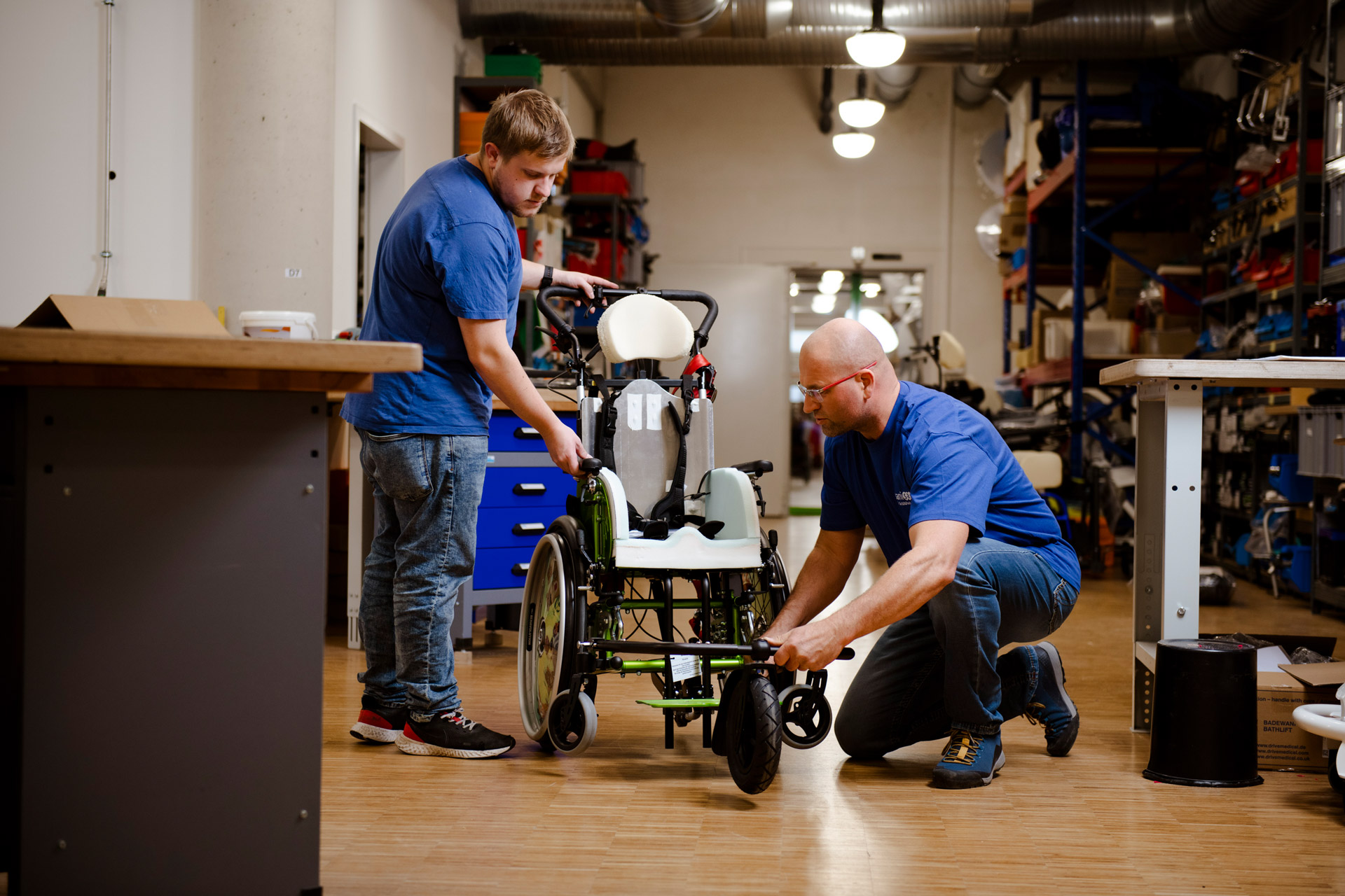 In einer Werkstatt arbeiten zwei Männer an der individuellen Anpassung eines Rollstuhls. Einer von ihnen kniet sich hin, um ein Vorderrad einzustellen.