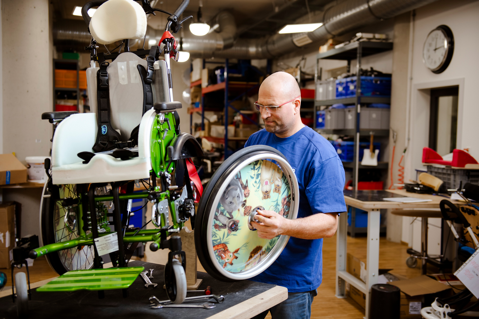 Ein Mann steht in einer Werkstatt und arbeitet an einem Kinderrollstuhl. Gerade passt er ein Rad an, welches bunt bedruckt ist.