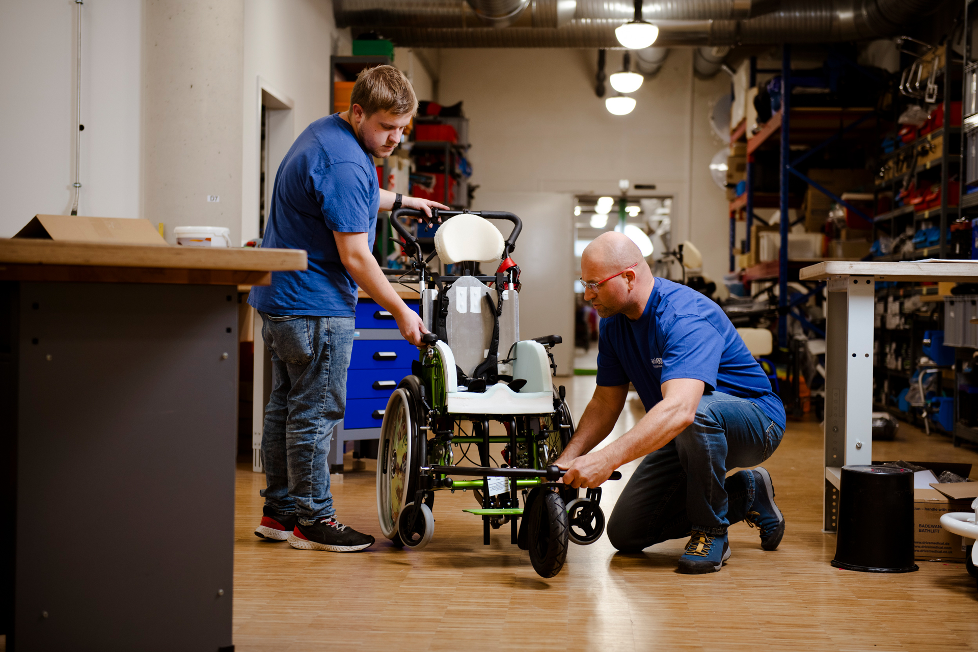 Zwei Männer arbeiten engagiert in einer Werkstatt an einem Rollstuhl, der individuell angefertigt wird. Einer von ihnen kniet nieder, um das Vorderrad anzupassen.