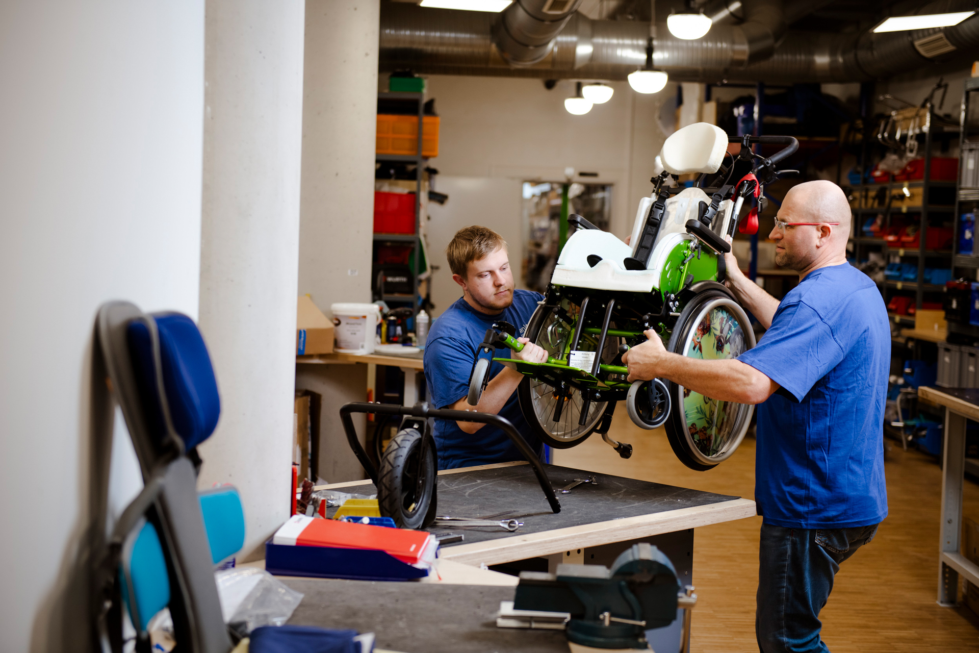 Zwei Männer arbeiten in einer Werkstatt an einem speziell angefertigten Rollstuhl. Sie heben den Rollstuhl gerade gemeinsam auf eine Werkbank.